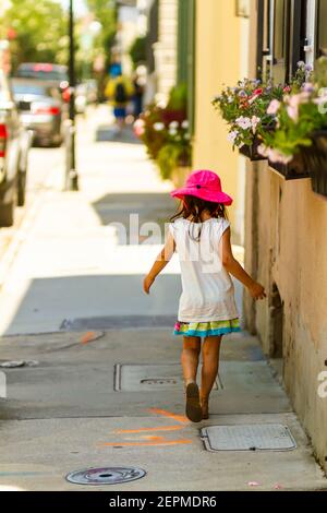 A pretty little girl wearing short summer dress, sandals and pink wide brimmed solar hat is walking alone in the shade at a sunny hot summer day. Sele Stock Photo