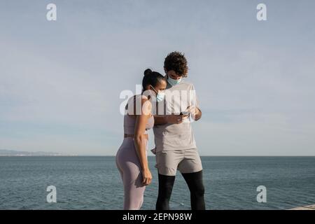 Unrecognizable ethnic sportsWoman in sterile mask near male partner surfing internet on cellphone against rippled sea under cloudy sky Stock Photo