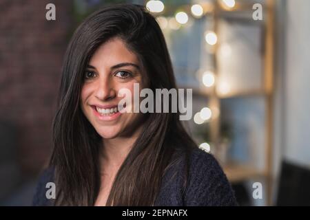 Cheerful female with dark hair looking at camera while standing in light room with brick wall and garland on shelf on blurred background Stock Photo