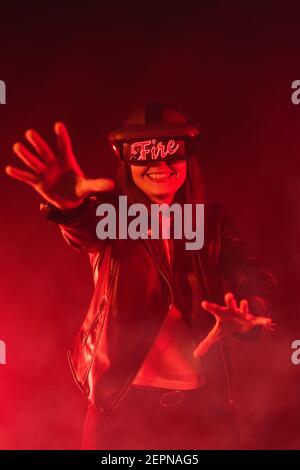 Anonymous female with outstretched arms wearing modern headset screaming while exploring cyberspace in room with red neon light and smoke Stock Photo