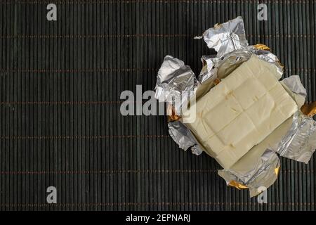 Rectangular piece of processed cheese in open foil package on a dark bamboo background. Cheese ready to eat. Dairy products, food. Stock Photo