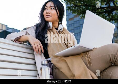 Ethnic young Asian female wearing warm outerwear browsing netbook while working remotely sitting on a bench on the street Stock Photo