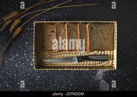 Top view of fresh homemade rye bread near knife in wicker basket and wheat spikes on table Stock Photo