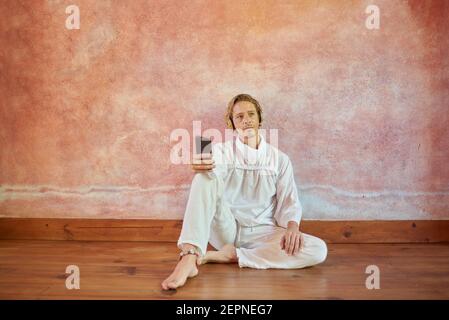Young mindful male in white outfit with cellphone sitting leaning on a wall listening to yoga course from headset looking away Stock Photo