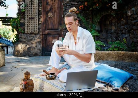 Young barefoot male text messaging on cellphone near netbook and Buddha statuette in patio after practicing yoga Stock Photo