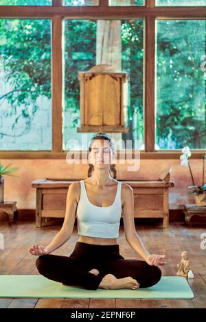 Young concentrated female with closed eyes practicing yoga with crossed legs near bowl gong in house Stock Photo