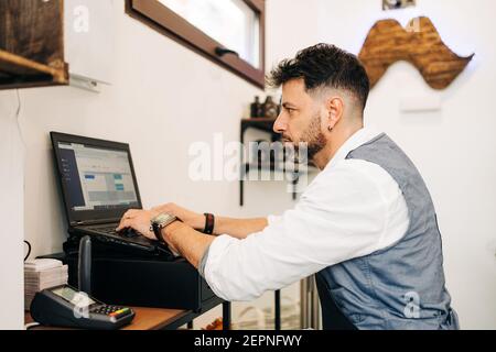 Side view of focused male barber working on netbook with open program on screen in beauty salon near cash register till and card reader Stock Photo