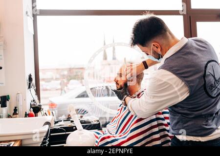 Back view of anonymous masculine stylist barber on face mask shaving beard of man using straight razor in beauty salon during coronavirus pandemic Stock Photo