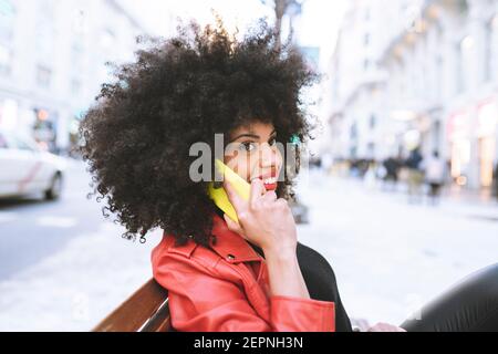 Side view of happy African American female with curly hair smiling widely while talking on smartphone on city street Stock Photo