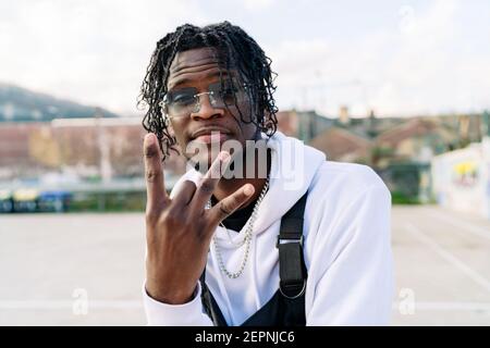 Trendy African American male in sunglasses with Afro braids demonstrating rap gesture while looking at camera in town Stock Photo