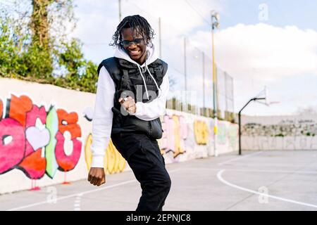 Young trendy ethnic man in sunglasses dancing on tiptoes while looking down against graffiti wall in city Stock Photo