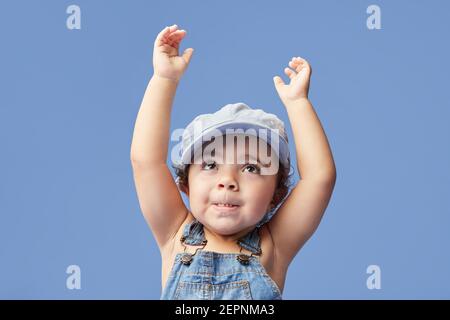Charming barefoot child in denim dress and dress with curly hair looking up with arms raised while dancing on blue background Stock Photo