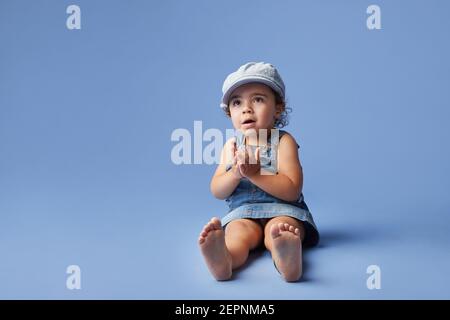 Charming cheerful barefoot child in denim dress and hat with curly hair looking away while playing on blue background Stock Photo