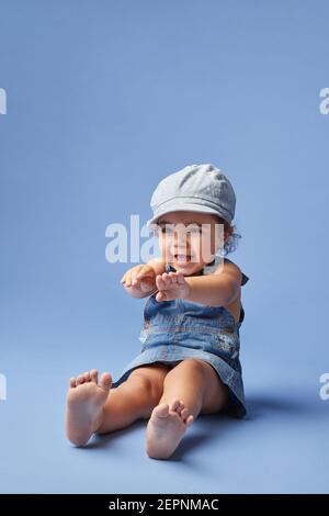 Charming cheerful barefoot child in denim dress and hat with curly hair looking away while playing on blue background Stock Photo