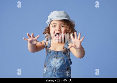 Charming barefoot child in denim dress and hat with curly hair looking a camera while standing on blue background making faces Stock Photo