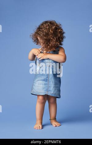 Charming barefoot child in denim dress with curly hair distracted with cloths while standing on blue background Stock Photo
