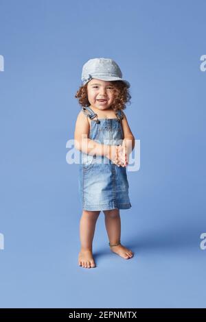 Charming cheerful barefoot child in denim dress and hat with curly hair looking at camera while dancing on blue background Stock Photo