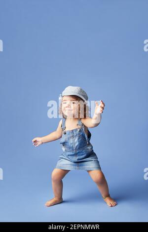 Charming barefoot child in denim dress and hat with curly hair looking away while dancing on blue background Stock Photo