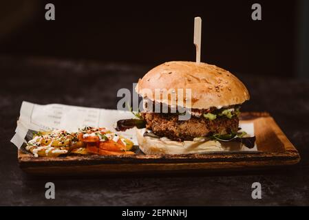 Yummy burger with vegetarian patty and grilled shiitakes between buns near sweet potato and carrot slices with alioli sauce on dark background Stock Photo