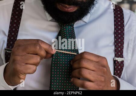 Crop unrecognizable ethnic boss wearing elegant white shirt and suspenders putting on tie lip while preparing for work Stock Photo