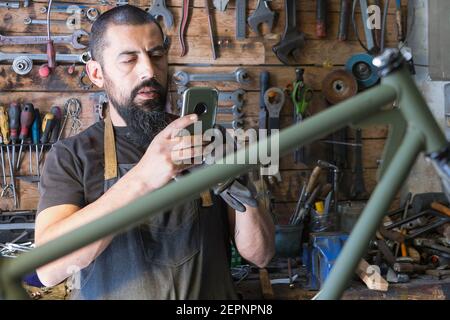 Concentrated bearded male master in apron taking photo of bicycle during repairing in garage Stock Photo