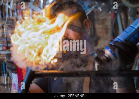 Serious bearded man in apron and protective gloves using professional equipment with fire for repair bicycle in workshop Stock Photo