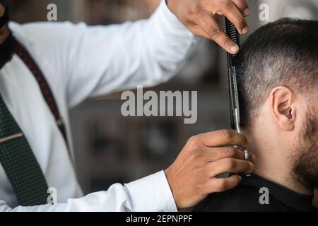 cropped unrecognizable ethnic male barber trimming hair of client with scissors during grooming procedure in beauty salon Stock Photo