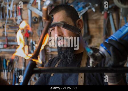 Serious bearded man in apron and protective gloves using professional equipment with fire for repair bicycle in workshop Stock Photo