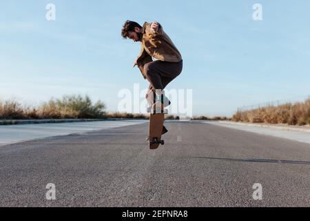 Full body young bearded skater in casual outfit jumping while performing kickflip on skateboard on asphalt road Stock Photo
