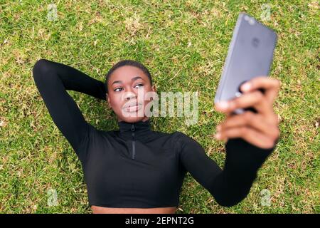 Young ethnic female in casual apparel with wireless headset taking self portrait on mobile phone lying in a park Stock Photo