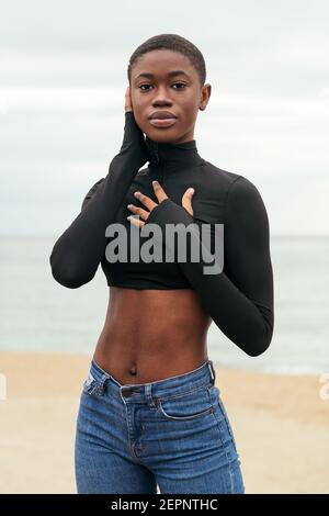 Young gentle African American female in casual clothes touching cheek while looking at camera on ocean coast Stock Photo