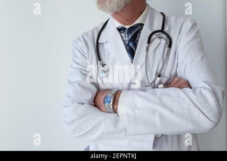 Crop senior male doctor with gray beard and folded arms in robe with stethoscope standing in clinic Stock Photo