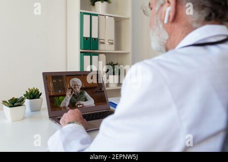 Back view of crop anonymous elderly male doctor in wireless earbud speaking with Woman on video call on netbook in clinic Stock Photo