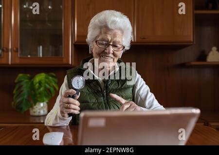 Smiling senior female pensioner pointing at sphygmomanometer while talking on video chat against tablet during online health consultation at home Stock Photo