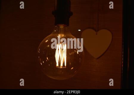 Glowing incandescent Edison light bulb closeup with heart in the background Stock Photo