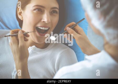 Smiling brunette woman being examined by dentist at dental clinic. Hands of a doctor holding dental instruments near patient's mouth. Healthy teeth Stock Photo