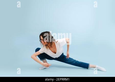 Professional contemporary dancer poses in front of blue studio background. Her legs in a low lunge, body lowered to the side. Arms bent in elbows, fac Stock Photo