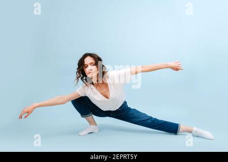 Bored contemporary dancer poses in front of blue studio background. Her legs in a low side lunge, body lowered, arms spread apart in a straight line. Stock Photo