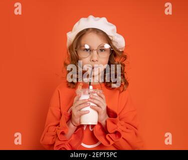 Beautiful little girl child 6-7 years in beret wearing glasses drinking milkshake through straw on bright orange background. Close up photo Stock Photo