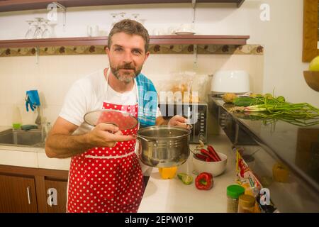 domestic lifestyle portrait of young attractive and relaxed home cook man in red apron cooking in kitchen preparing delicious and healthy food Stock Photo