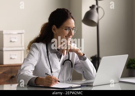 Smiling professional female doctor taking notes, looking at laptop screen Stock Photo