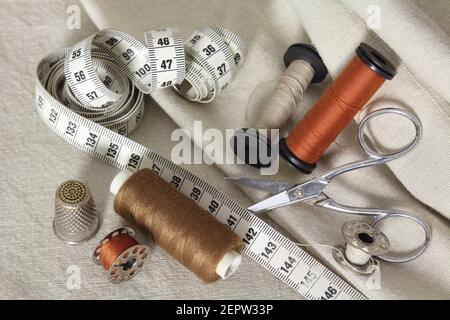 Set of sewing accessories: spools of brown thread, sepia and a needle, a pair of scissors and a tape measure. Tailor Accessories laid on beige fabrics Stock Photo
