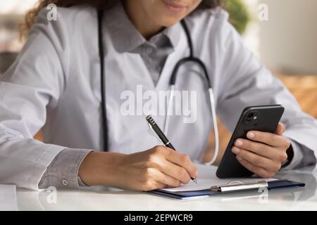 Close up female doctor wearing uniform holding smartphone, taking notes Stock Photo