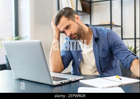 Young tired bearded office worker freelancer in casual clothes sitting at the desk at his workplace, supporting head with hand and looking at the laptop screen, feeling overworked and sleepy Stock Photo