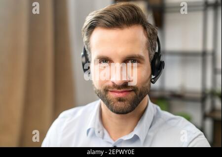 Positive young handsome bearded man employee in headset with microphone, working in the customer service department, looking at the camera, talking to customers on a helpline at the call center Stock Photo