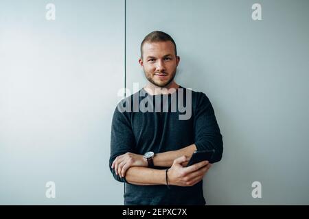 Portrait of a handsome young businessman standing with mobile phone. Male professional in casuals looking at camera against a white wall. Stock Photo