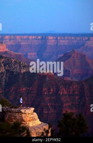 A young man stands on the edge of the Grand Canyon at sunset.