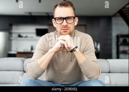 Portrait of handsome young bearded hipster man with his hands on chin looking at camera, wearing glasses and wireless earphones, sitting on a couch in the living room. The view while video-calling Stock Photo