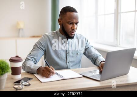 Black Businessman Working Taking Notes And Using Laptop At Workplace Stock Photo