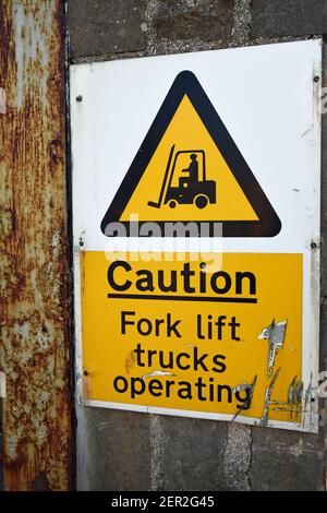 Caution fork lift trucks operating sign against industrial wall background. Yellow warning triangle with icon, text. Stock Photo